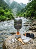 TrailChef™ 10-Piece Camping Cookware Set placed on a flat stone next to a flowing river in a lush green valley